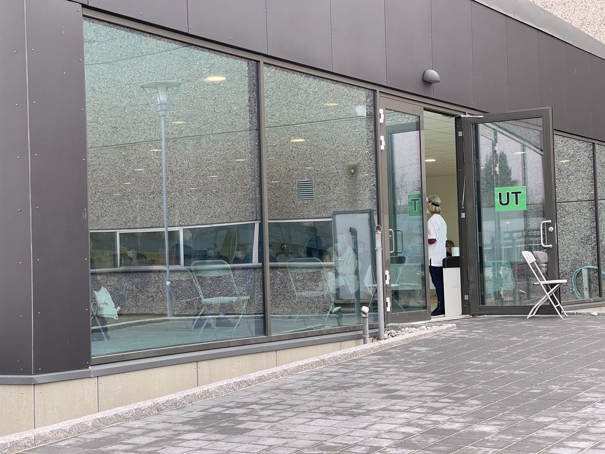 Mennesker med munnbind venter innenfor glassveggen på vaksinesenteret